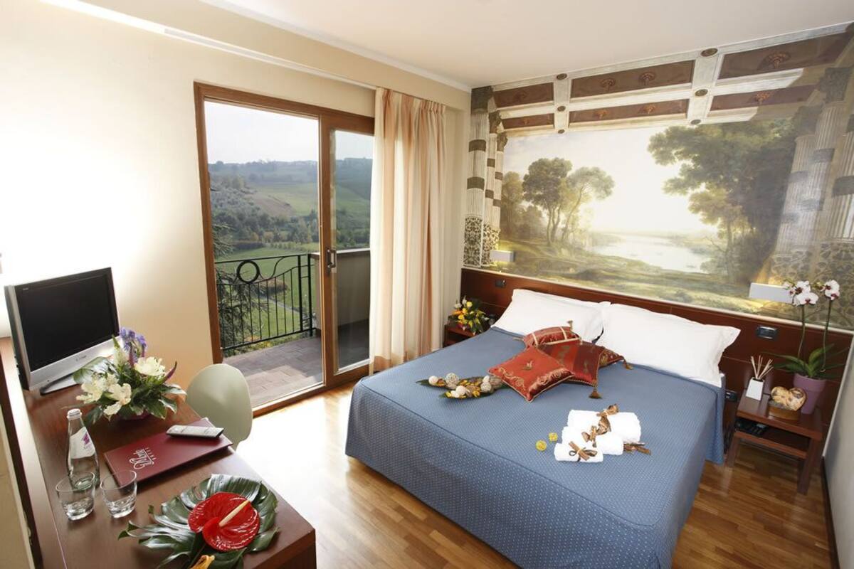 A hotel room with a bed, a desk with a computer, a window with a view of the countryside, and a painting on the wall.