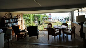 Una habitación iluminada por el sol, con un ventanal grande, una estantería, una mesa redonda con sillas y vistas a una playa y un faro.