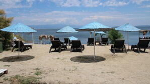 Una playa con varias sillas de playa y sombrillas azules, dispuestas en una zona arenosa con vista al mar y montañas distantes.