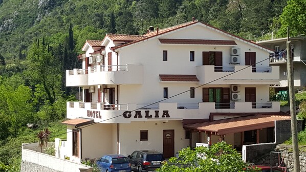 A two-story hotel named "GALIA" with a white facade, brown shutters, and balconies. There are cars parked in front, and the hotel is surrounded by greenery and hills. - Hotel Galia (Kotor)