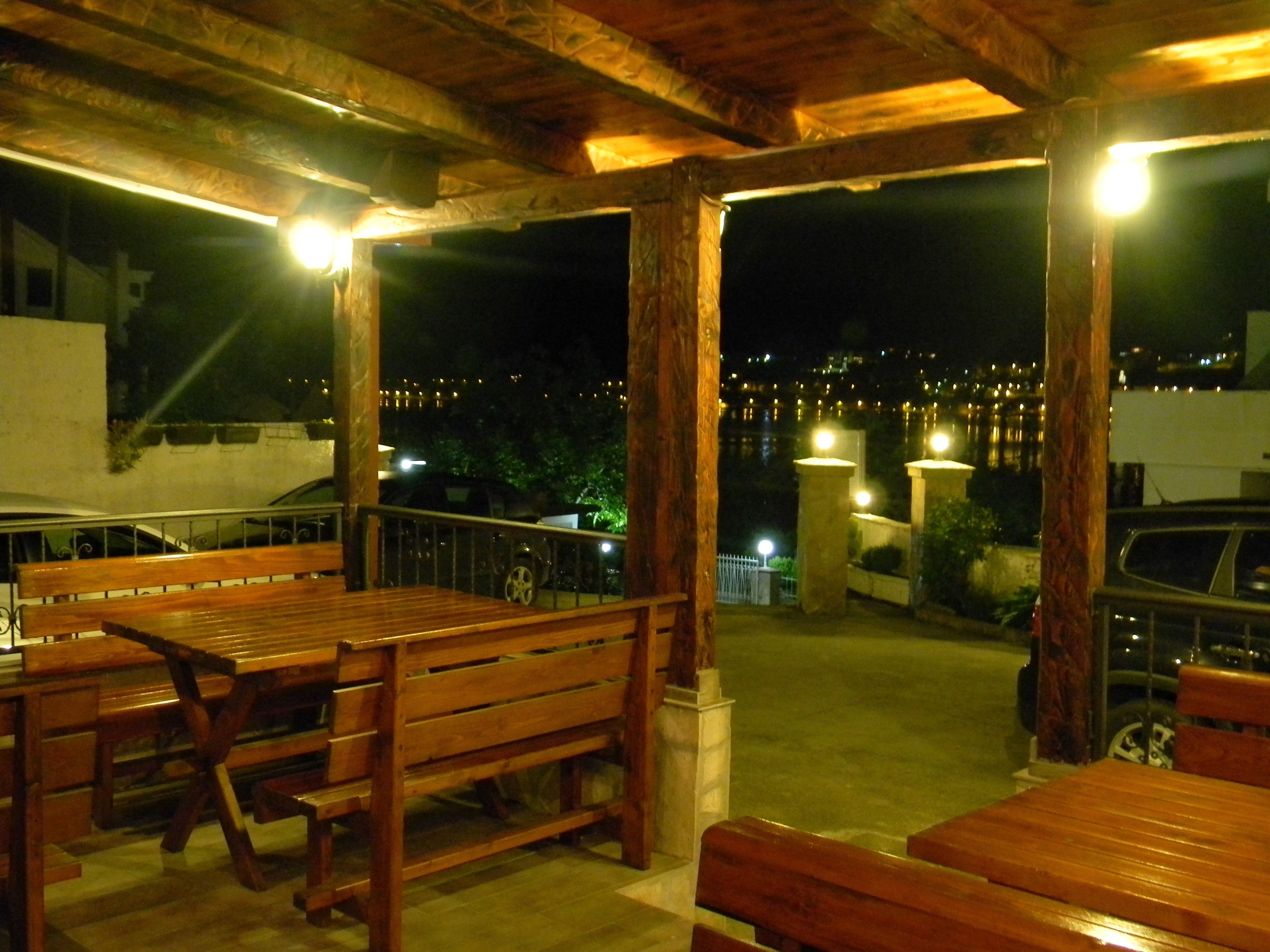 Un patio exterior con bancos y mesas de madera, barandilla y vista panorámica de la ciudad de noche.