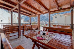 An outdoor dining area with wooden benches and tables, stone walls, and a view of mountains and a town. - Hotel Galia (Kotor)