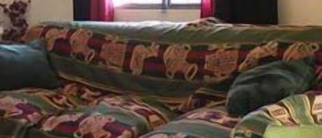 Um sofá verde com capa estampada colorida em um quarto com janela e cortinas.