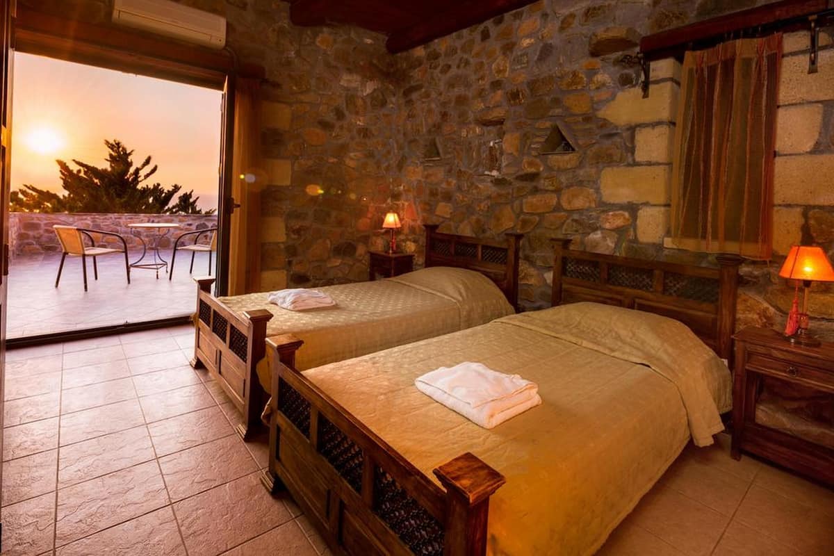 A room with two beds, stone walls, a balcony with chairs, and a sunset view.