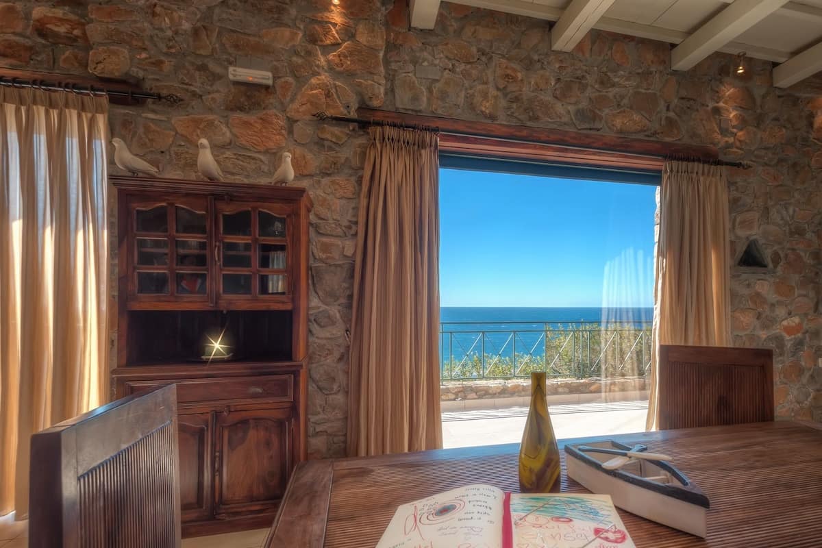 A room with a stone wall, a wooden cabinet, a table with a book and a bottle, and a view of the sea.