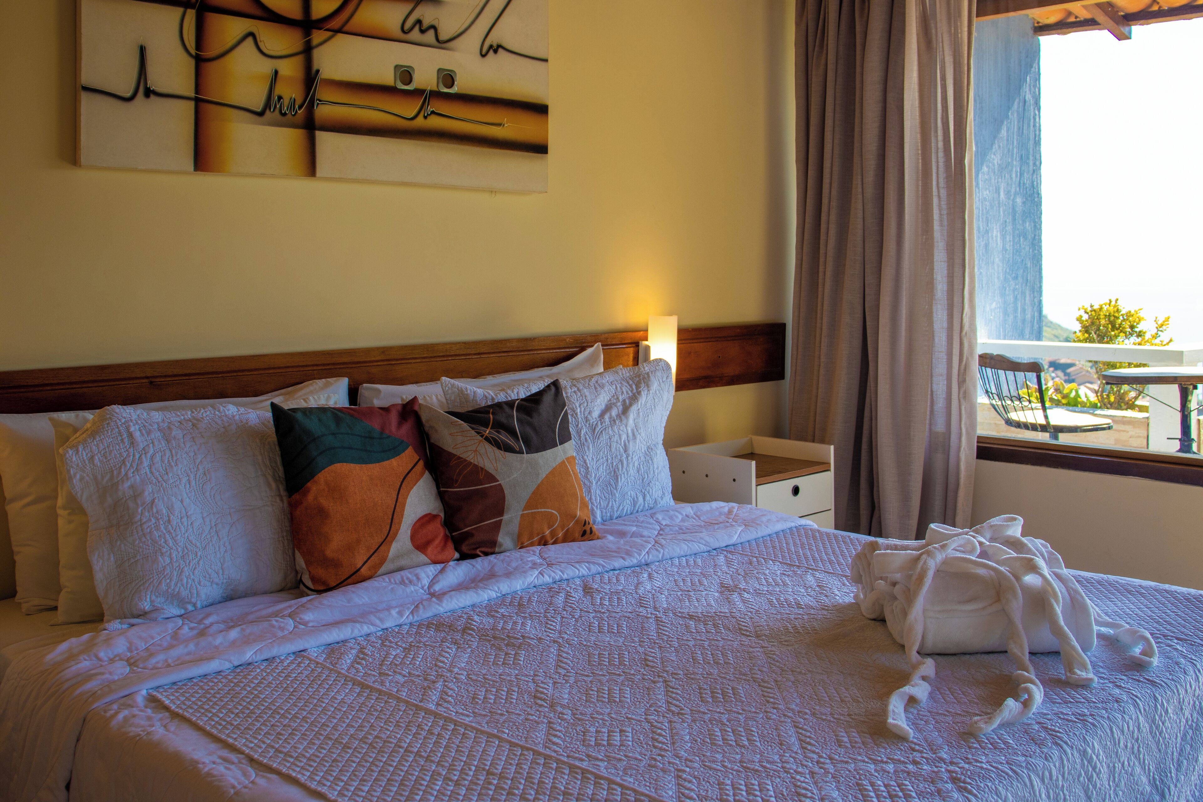 A neatly made bed with a variety of pillows, a bedside table with a lamp, and a window with curtains.