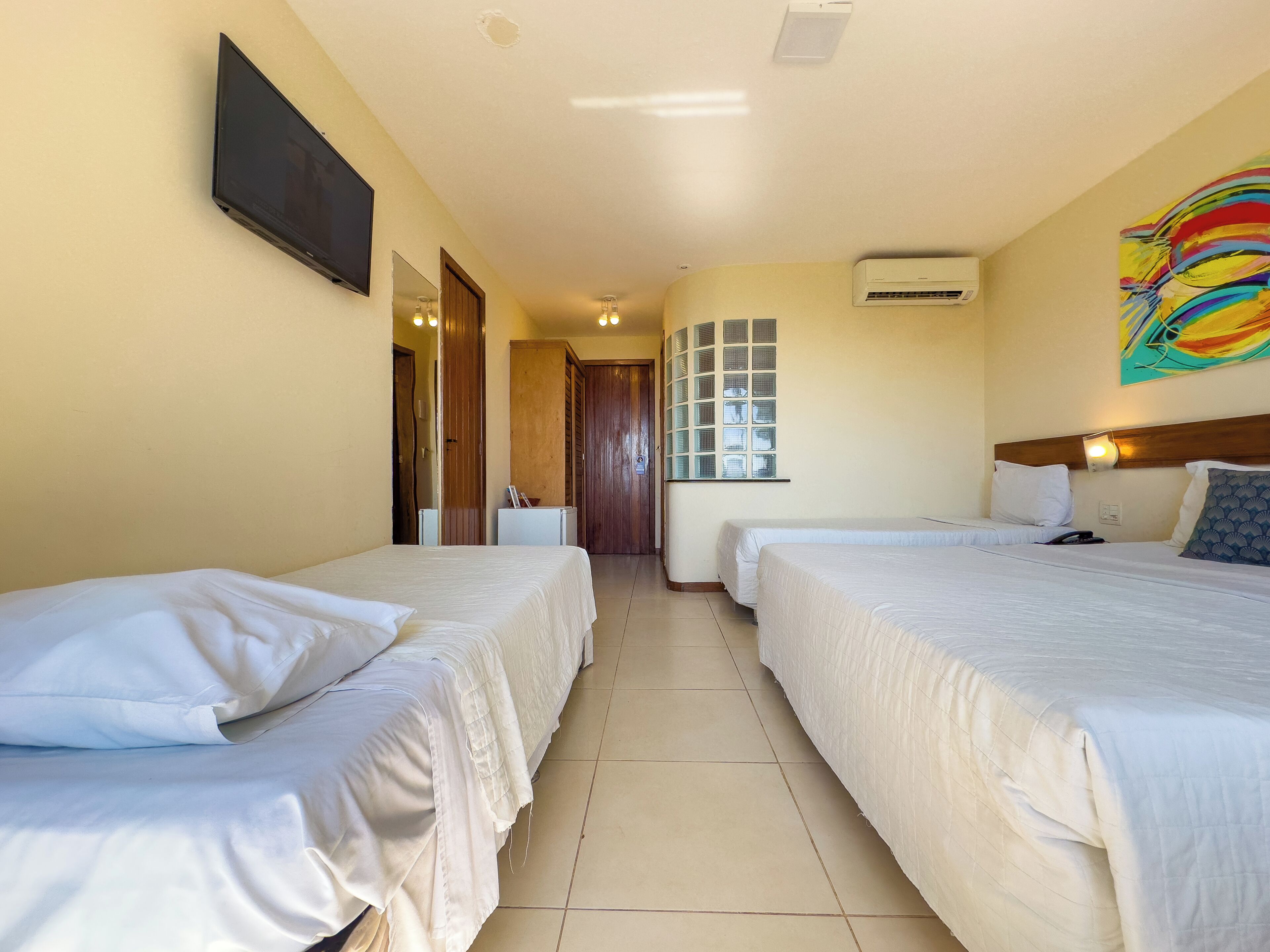 A hotel room with two beds, a TV mounted on the wall, an air conditioning unit, and a colorful abstract painting.