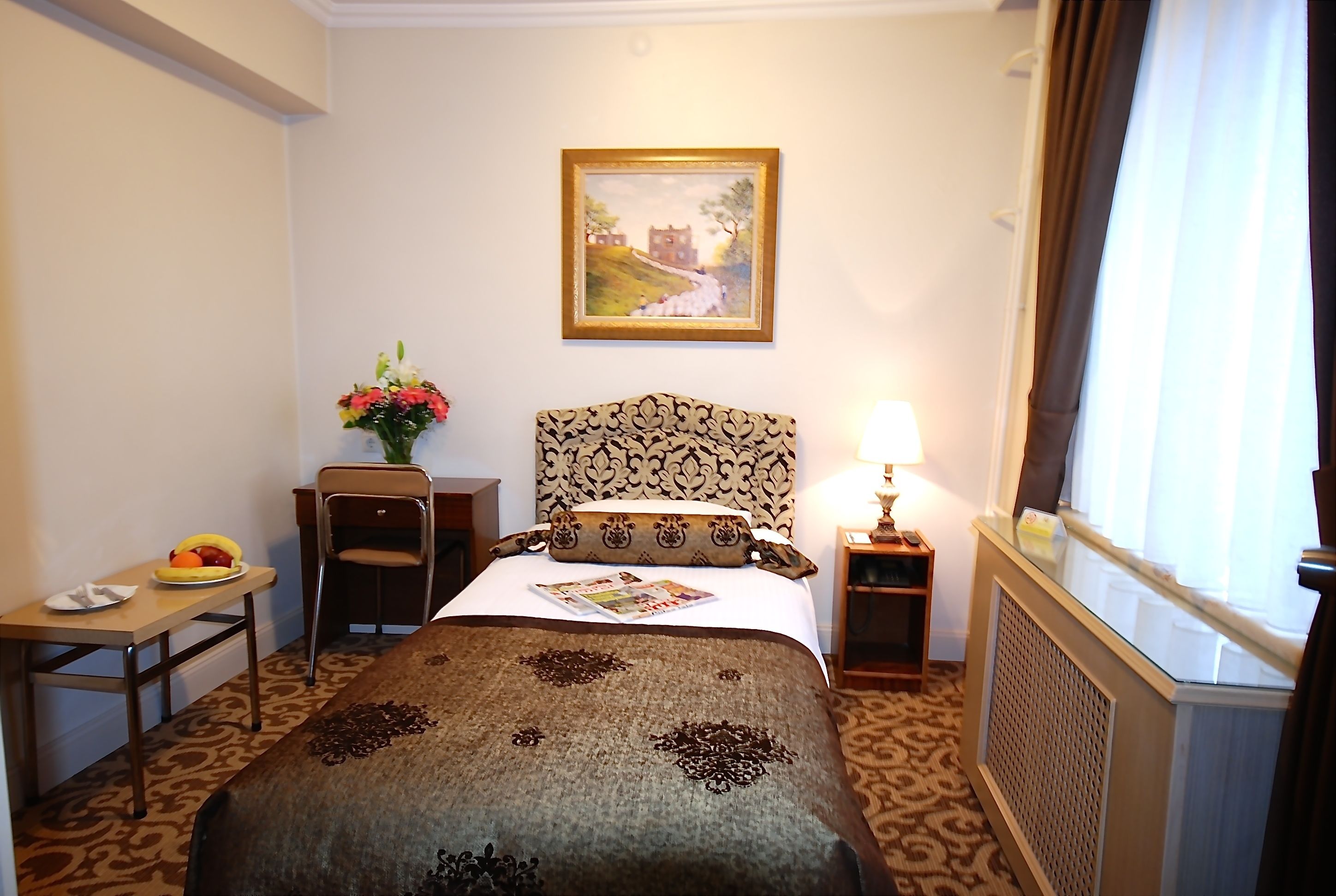 A hotel room with a bed, a desk, a chair, a lamp, a painting on the wall, and a window with curtains.