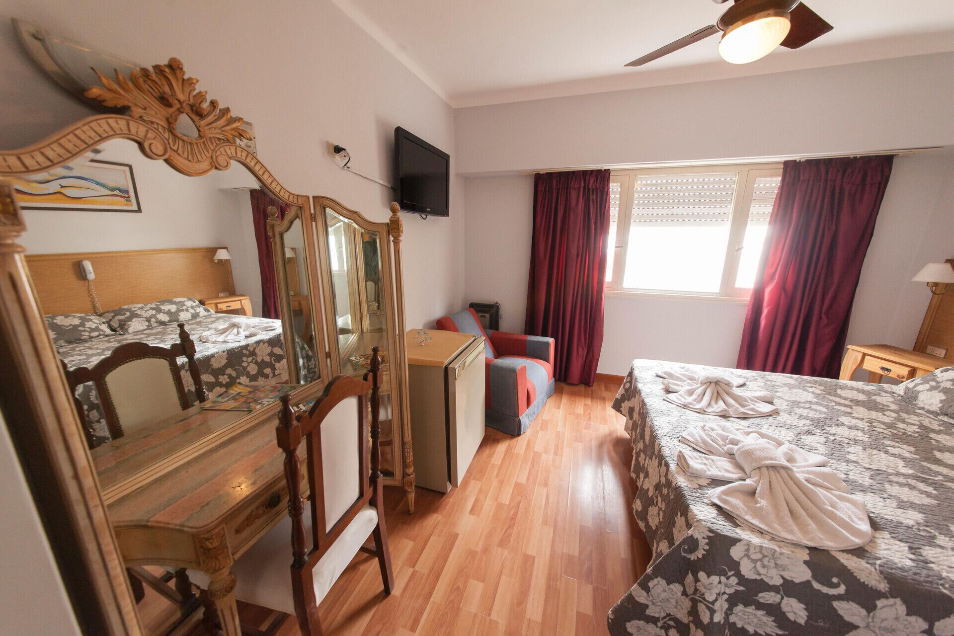 A hotel room with a wooden bed, a large mirror, a desk with a chair, a television, and a window with curtains.