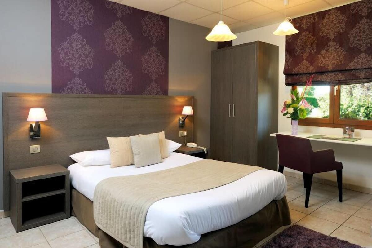 A hotel room with a large bed, bedside lamps, a desk, and a window with a view of greenery.