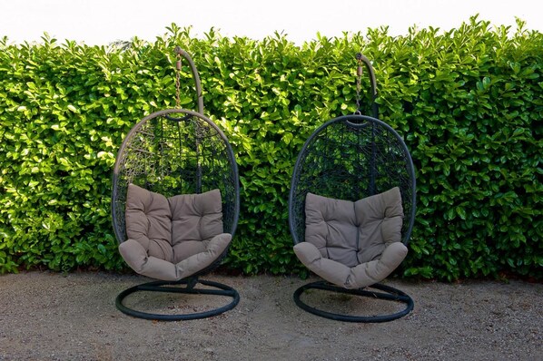 Dos sillones colgantes tipo huevo con cojines, sobre un fondo de seto.