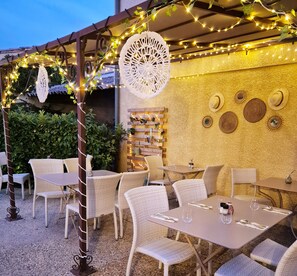 An outdoor patio with wicker furniture, string lights, and decorative wall hangings. - Logis Hôtel Le Mistral - Restaurant - Montélimar sud (Chateauneuf-du-Rhone)
