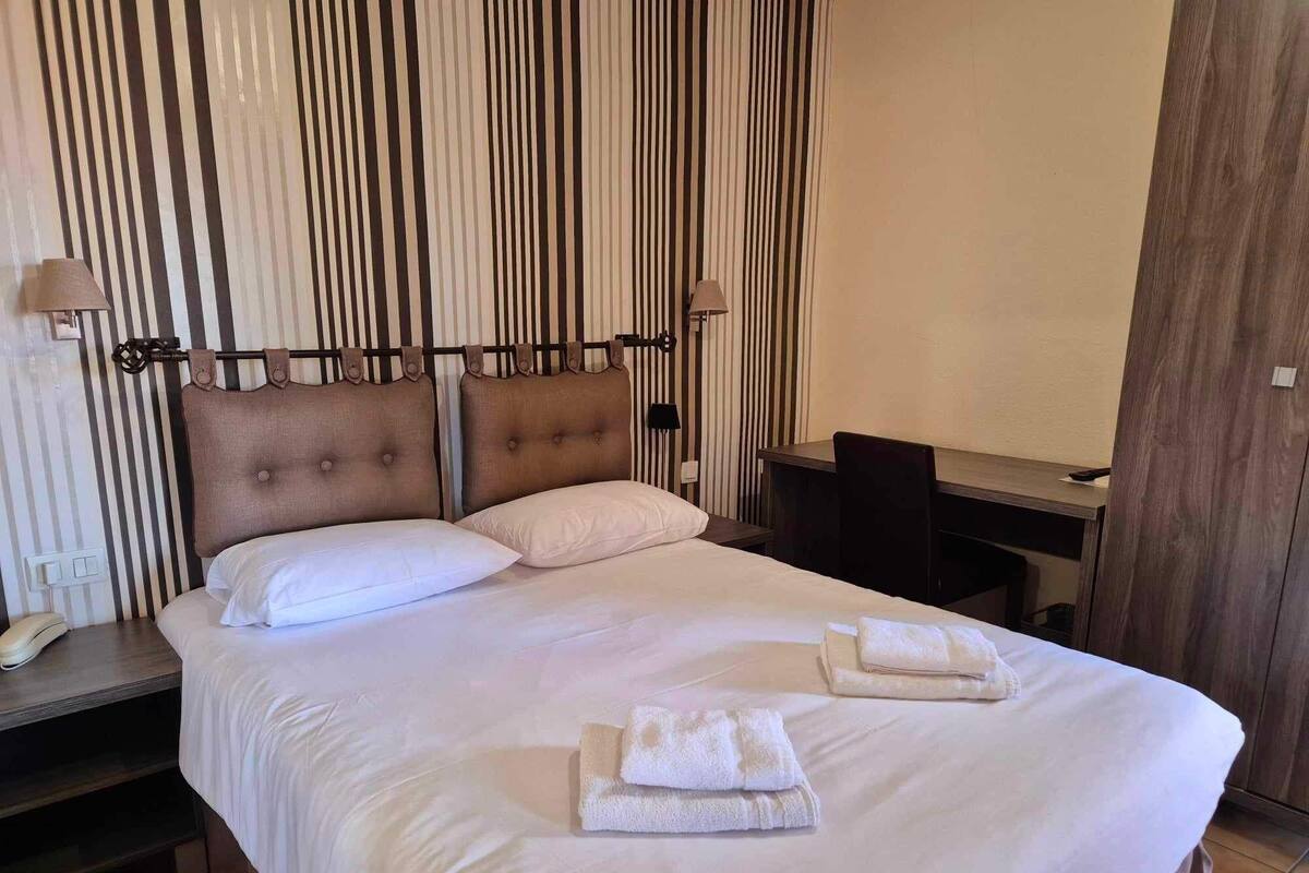 A neatly made bed with two pillows and two folded towels, a bedside table with a lamp, and a wooden desk with a chair.
