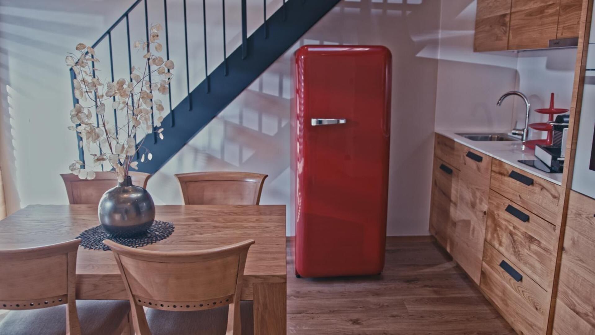 Une cuisine moderne avec un réfrigérateur rouge, des meubles de rangement en bois, une table à manger avec des chaises et un vase contenant des fleurs séchées.