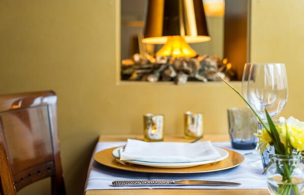A dining table set with a white plate, gold charger, and cutlery, a vase with yellow flowers, and a lit lamp in the background. - Els Caçadors (Ribes De Freser)