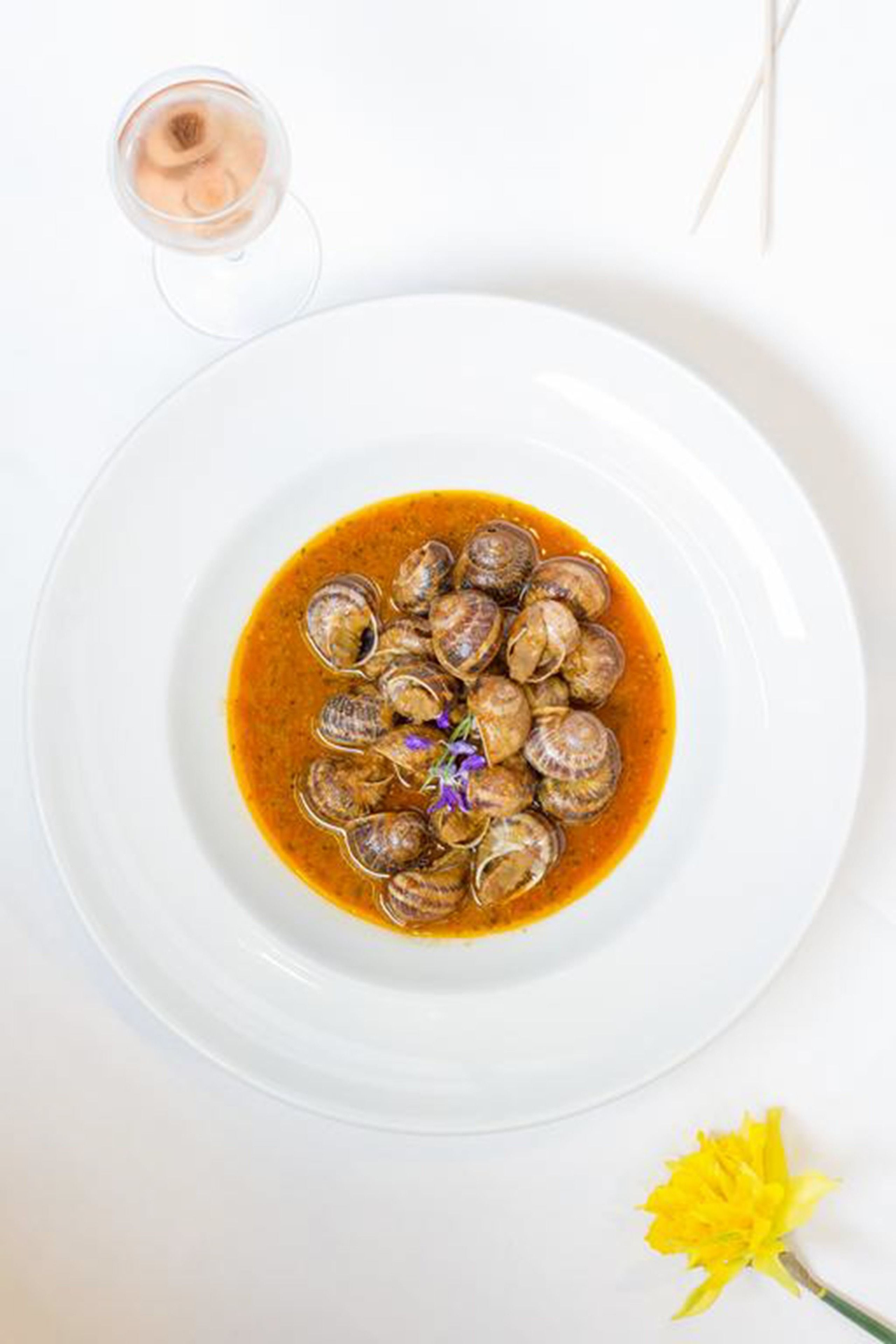 Un plato blanco con almejas en salsa marrón, una copa de vino blanco y una flor amarilla sobre un fondo blanco.