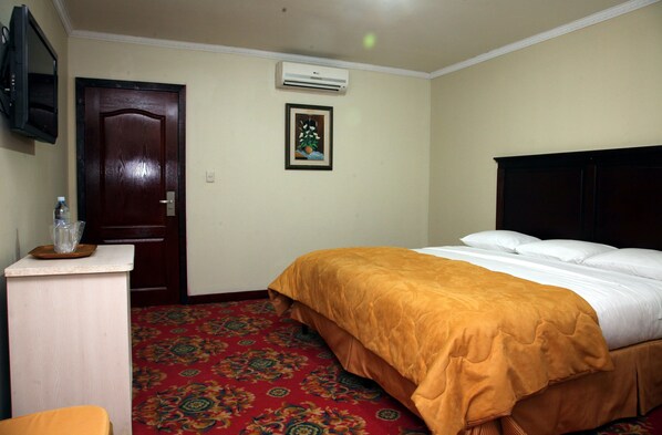 A hotel room with a bed, a wooden door, an air conditioning unit, a framed picture on the wall, and a carpeted floor.