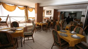 Une salle à manger avec plusieurs tables et chaises, un serveur en uniforme et une vue sur l’extérieur grâce à de grandes fenêtres.
