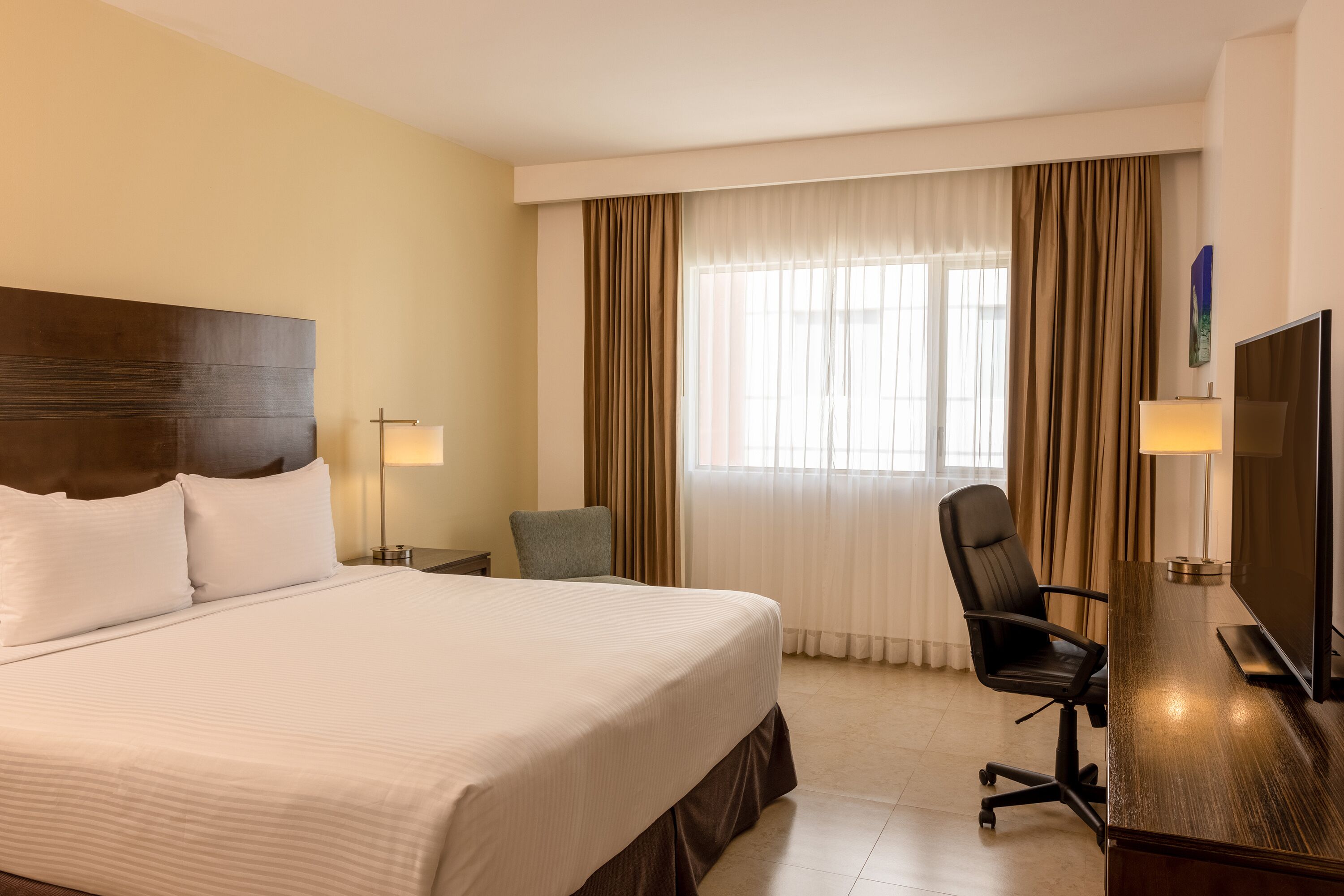 A hotel room with a large bed, a desk with a TV, a chair, and two lamps.
