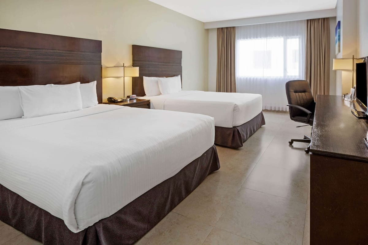 Habitación de hotel con dos camas, un escritorio, una silla y un ventanal con cortinas.