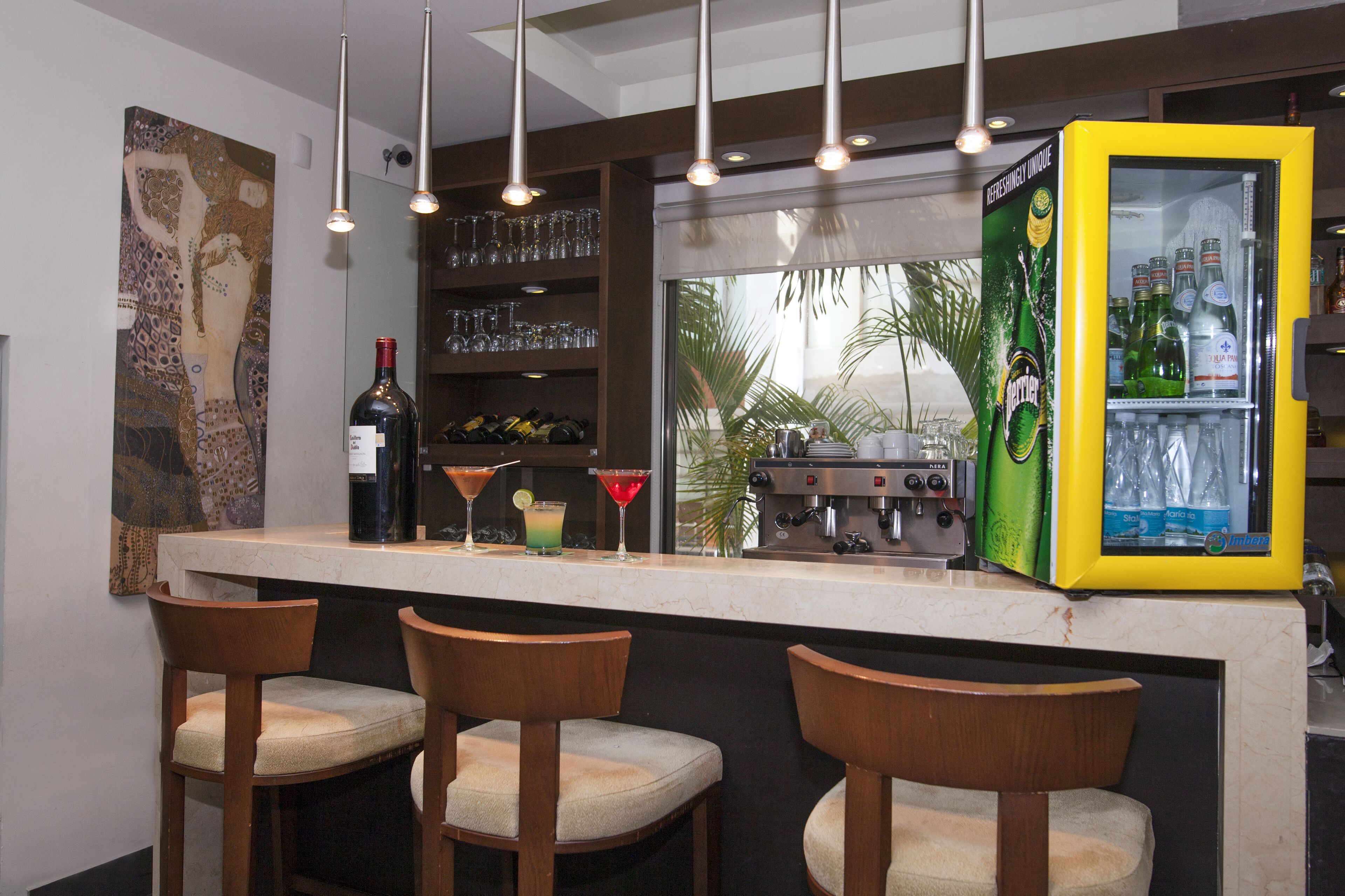 A bar area with a counter, three wooden bar stools, a refrigerator stocked with beverages, a coffee machine, and a large window with a view of palm trees.