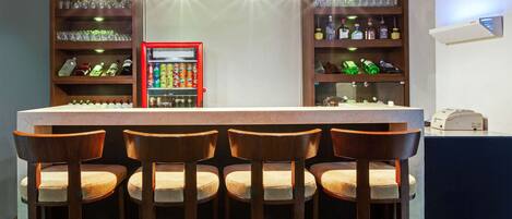Uma área de bar com balcão, quatro banquetas de madeira, prateleiras com garrafas e copos, e um dispensador de bebidas vermelho.