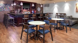 Eine Bar mit Backsteinwand, mehreren Barhocker und einem runden Tisch mit Stühlen. Im Hintergrund befindet sich eine Couch und ein kleiner Tisch mit Stühlen.
