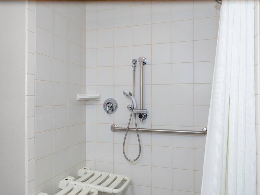 Área de duche com barra de apoio, chuveiro manual e parede revestida a azulejos brancos.