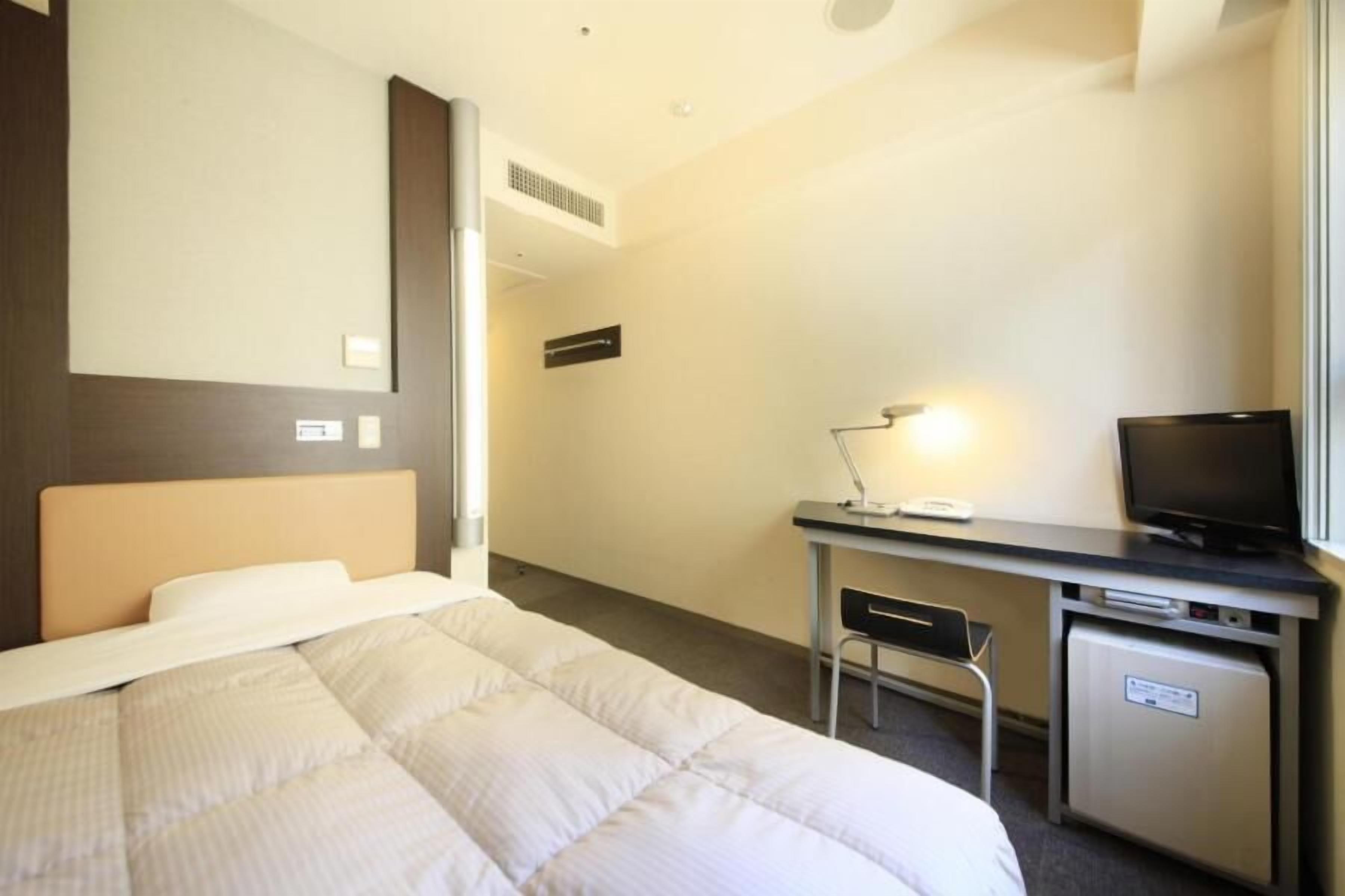 Una habitación de hotel con cama, escritorio, silla, televisor y un refrigerador pequeño.