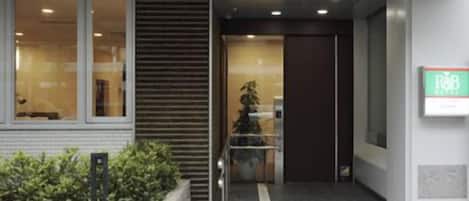 A modern building entrance with a glass door, a small garden area with plants, and a sign on the wall.