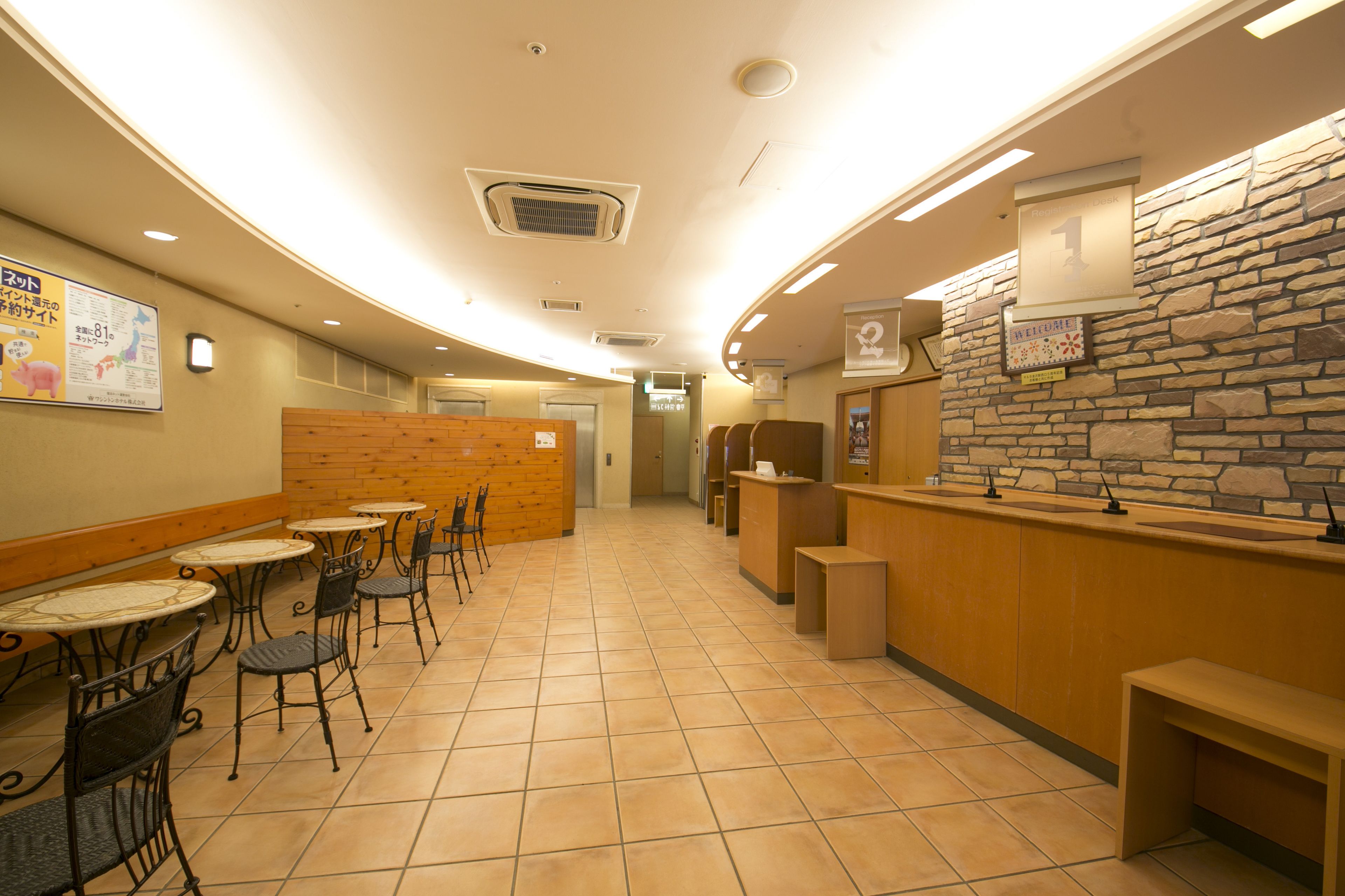 A hotel lobby with a curved ceiling, tiled floor, and a reception desk with a stone wall backdrop. There are tables and chairs arranged for guests.