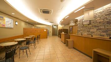 A hotel lobby with a curved ceiling, tiled floor, and a reception desk with a stone wall backdrop. There are tables and chairs arranged for guests.