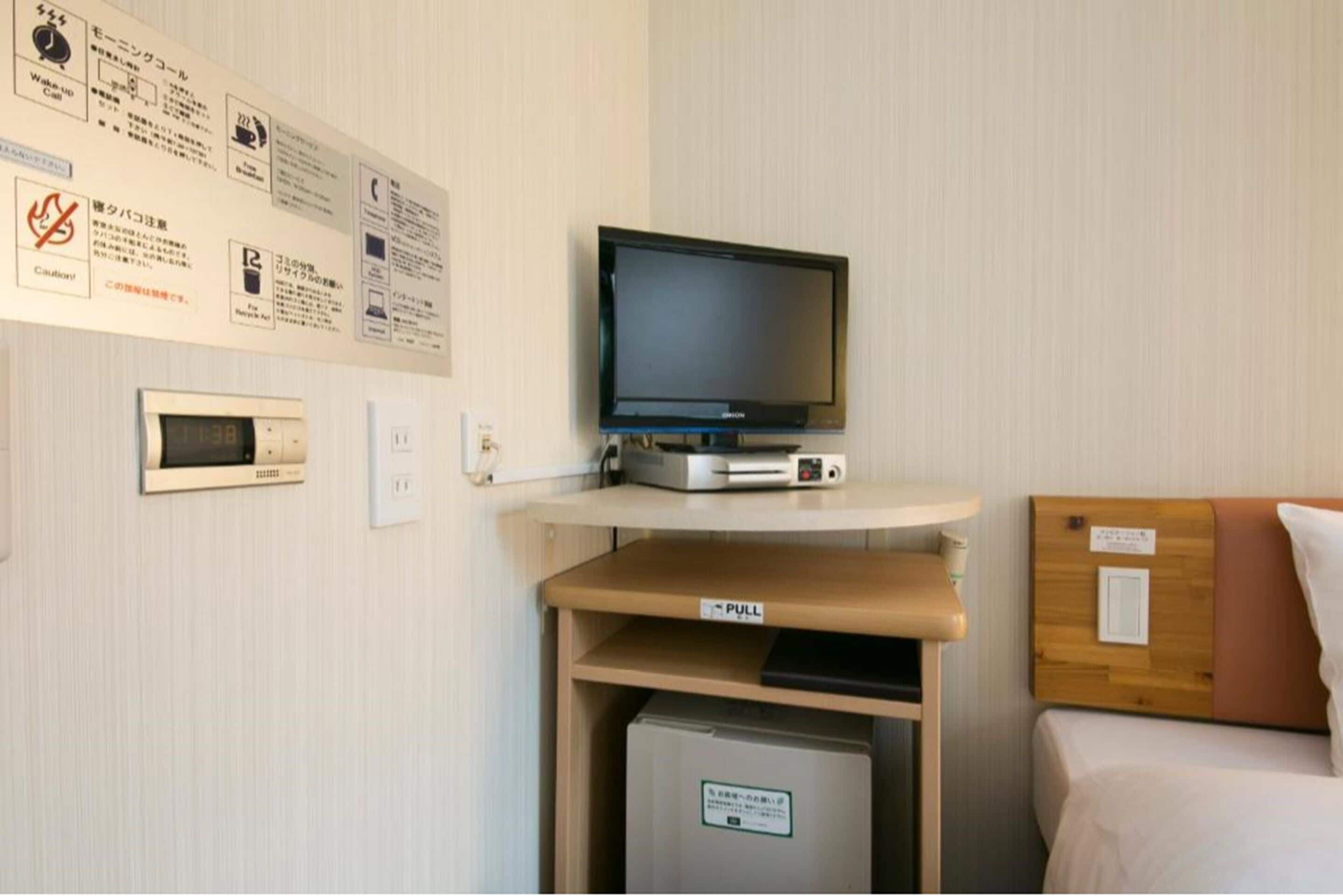 A hotel room with a television on a stand, a small refrigerator, a pull-out bed, and various informational signs on the wall.