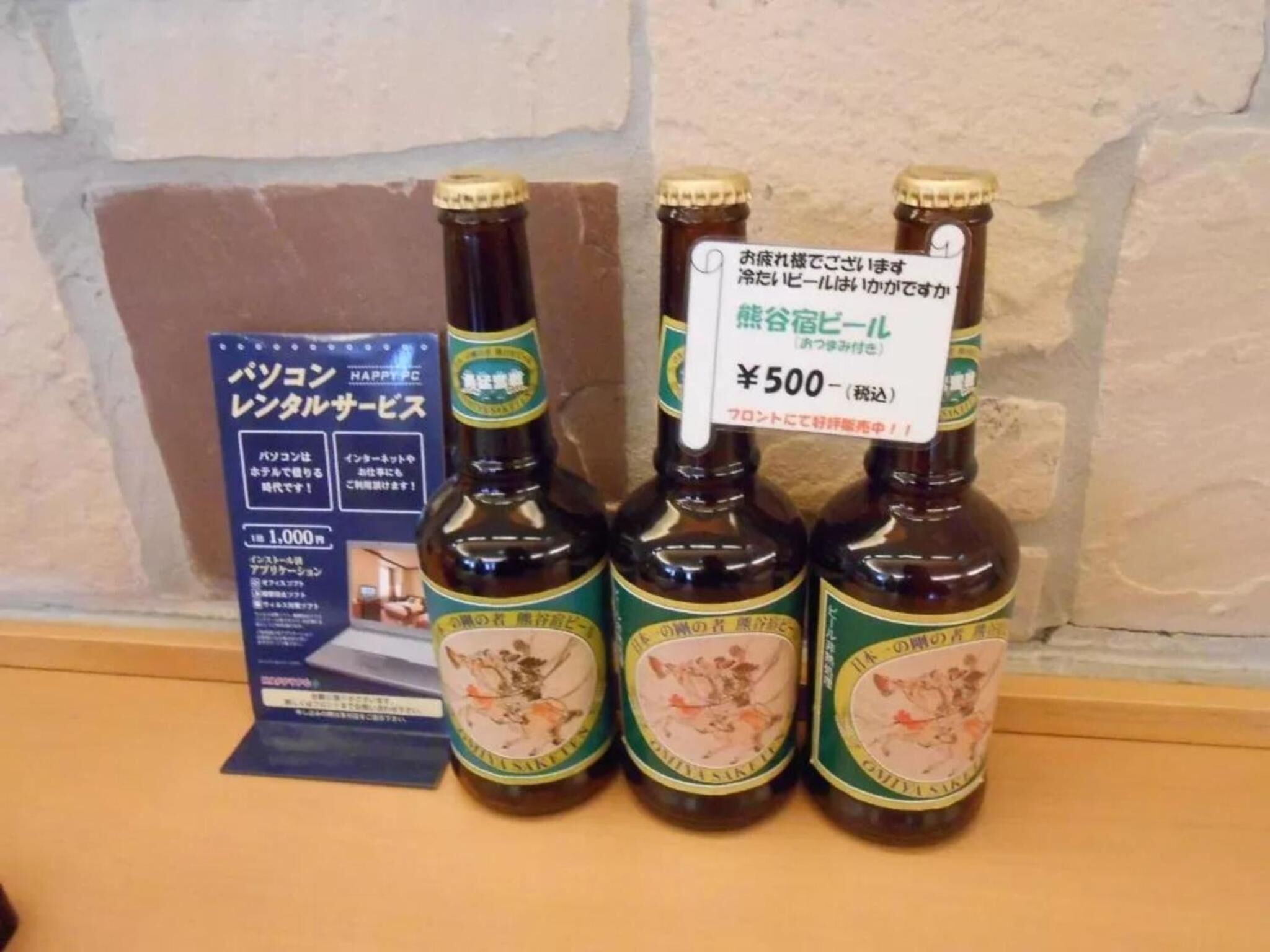 Three bottles of beer with labels, a price tag, and a promotional poster on a shelf.