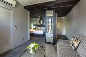A modern hotel room with a bed, a sofa, a coffee table with flowers, a mirror, and an air conditioning unit. - DoubleTree by Hilton Queenstown (Kawarau Falls)