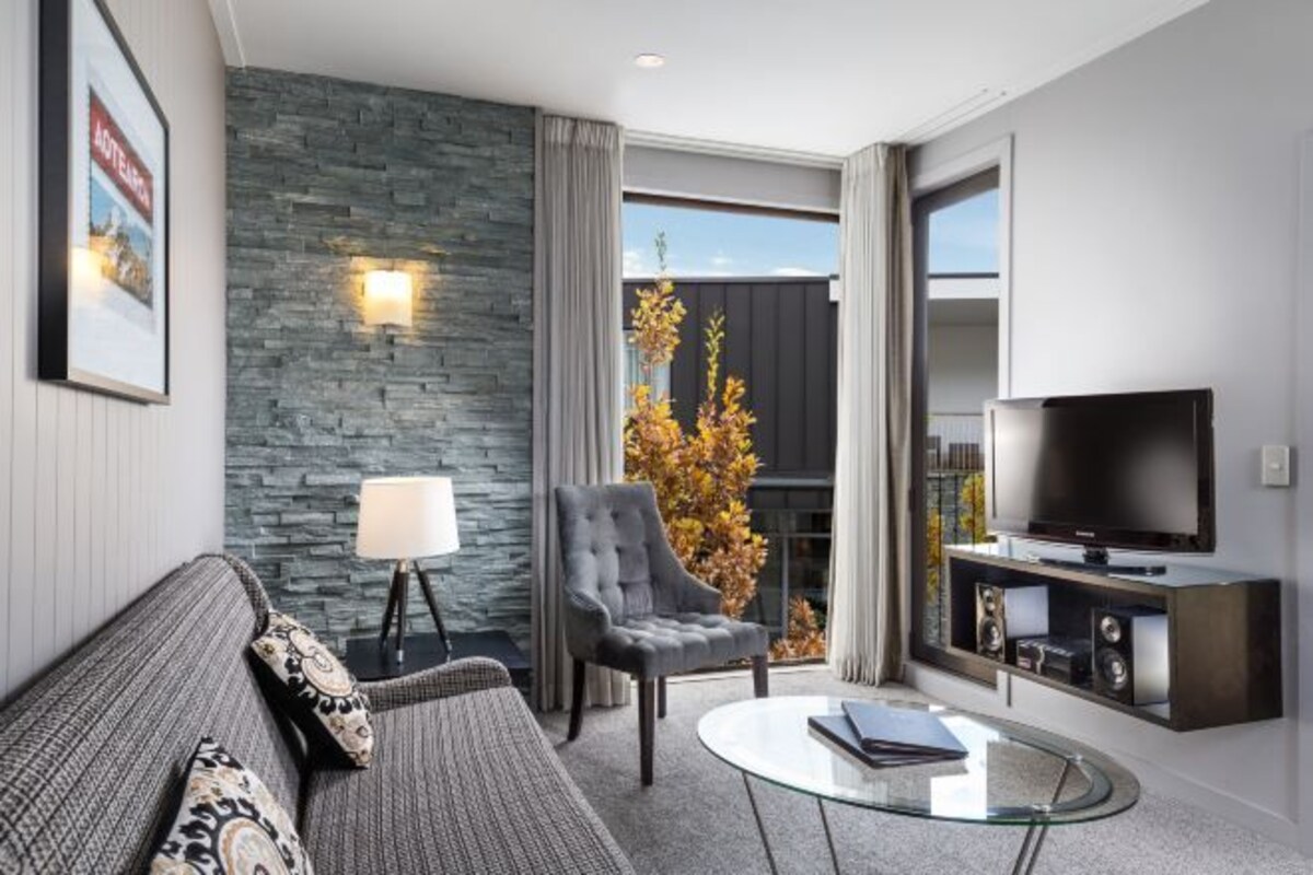 Una sala moderna con una pared de acento de piedra, un sofá gris, una mesa de centro de vidrio, un sillón gris, un televisor sobre un mueble de madera oscura y vistas de follaje otoñal a través de amplias ventanas.