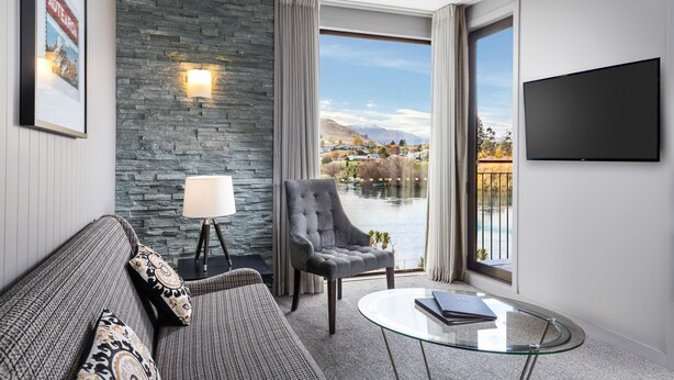 A modern living room with a stone wall, a sofa, a chair, a glass coffee table, and a TV mounted on the wall. - DoubleTree by Hilton Queenstown (Kawarau Falls)