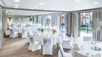 Une salle de banquet spacieuse, dotée de tables rondes, de nappes blanches et de chaises, avec de grandes fenêtres offrant une vue sur l’extérieur.