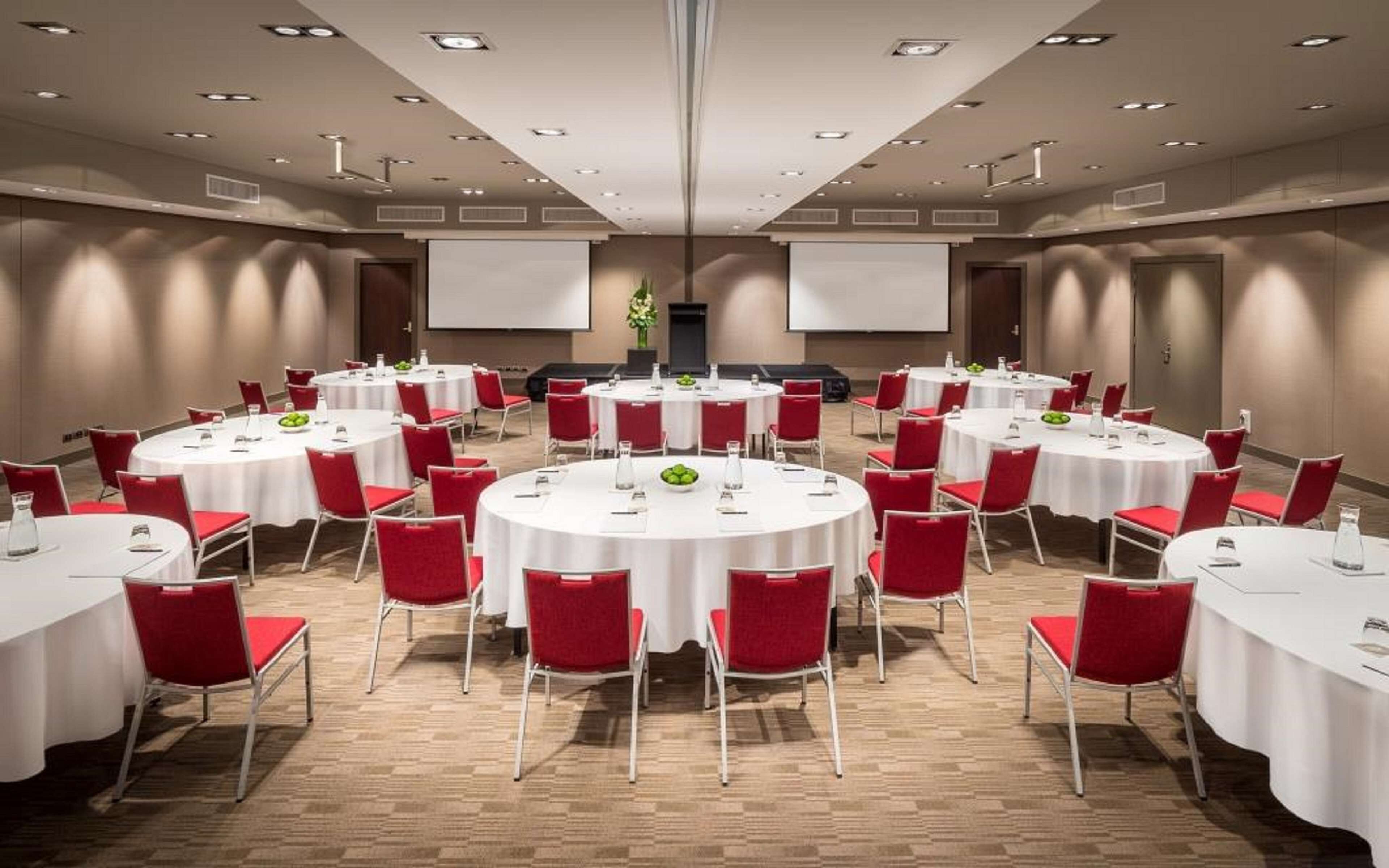 A conference room with multiple round tables, each with white tablecloths and red chairs, equipped with nameplates and water bottles.