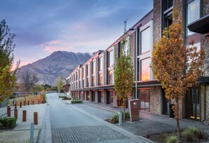 Un edificio moderno con fachada de piedra, ventanas amplias y vista a la calle con montañas al fondo.