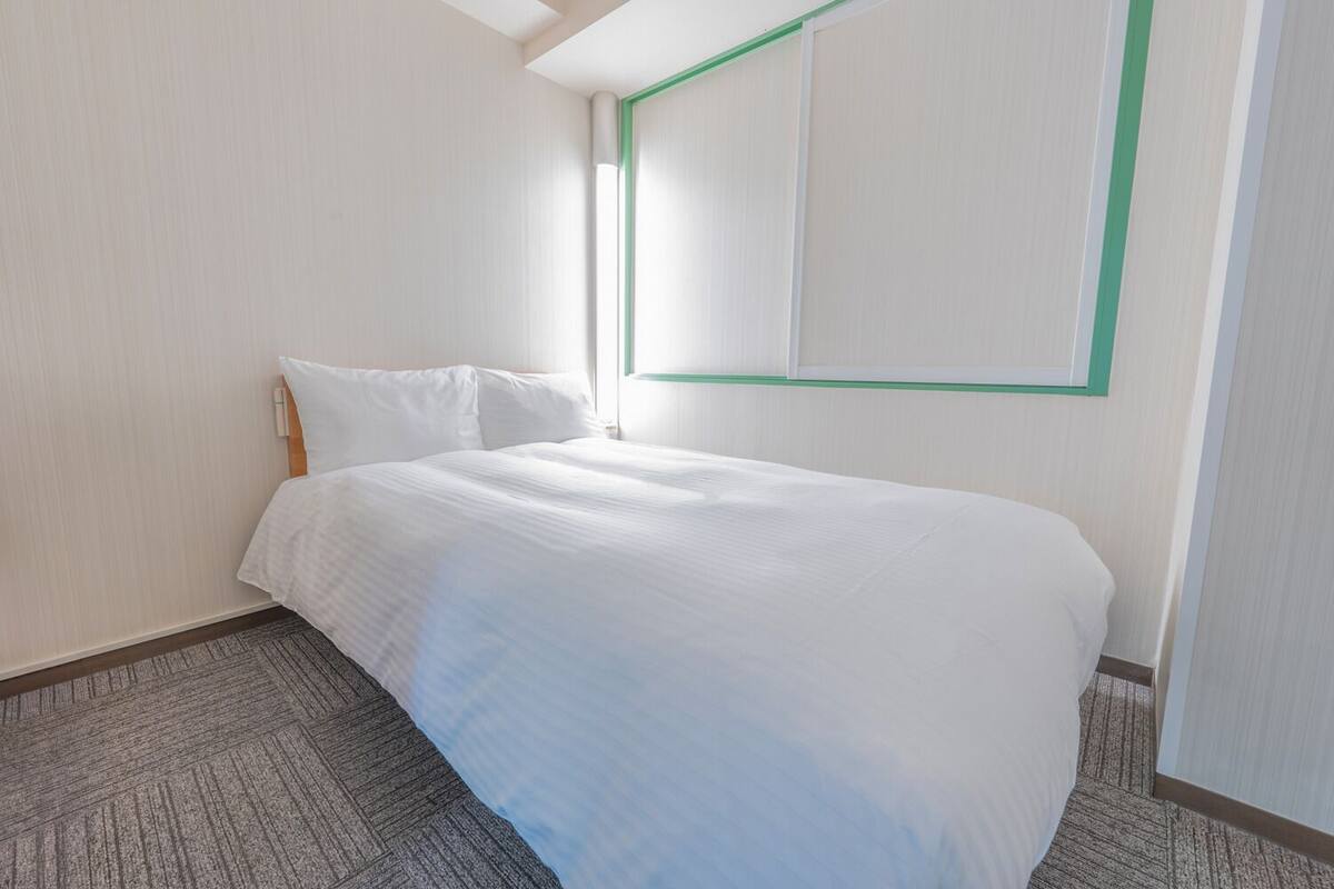 A hotel room with a neatly made bed, a window with a green frame, and a carpeted floor.