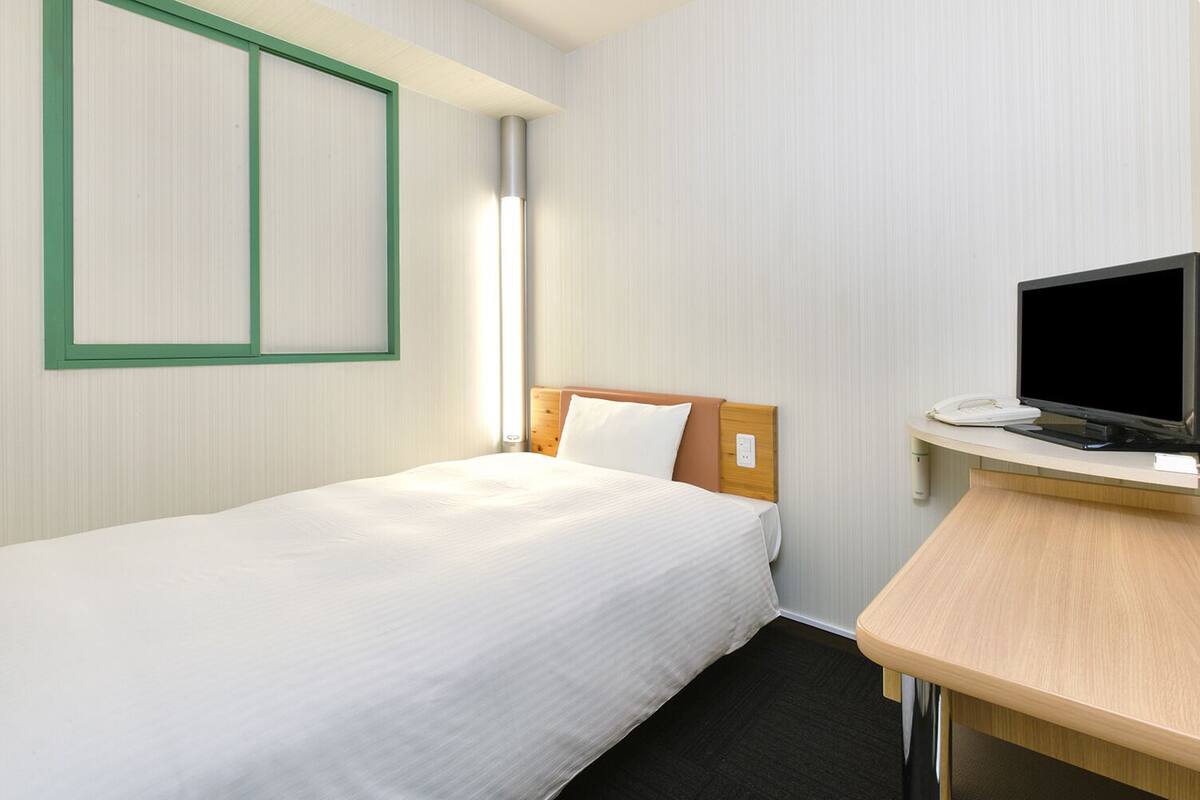 A hotel room with a single bed, a desk with a flat-screen TV, and a telephone.