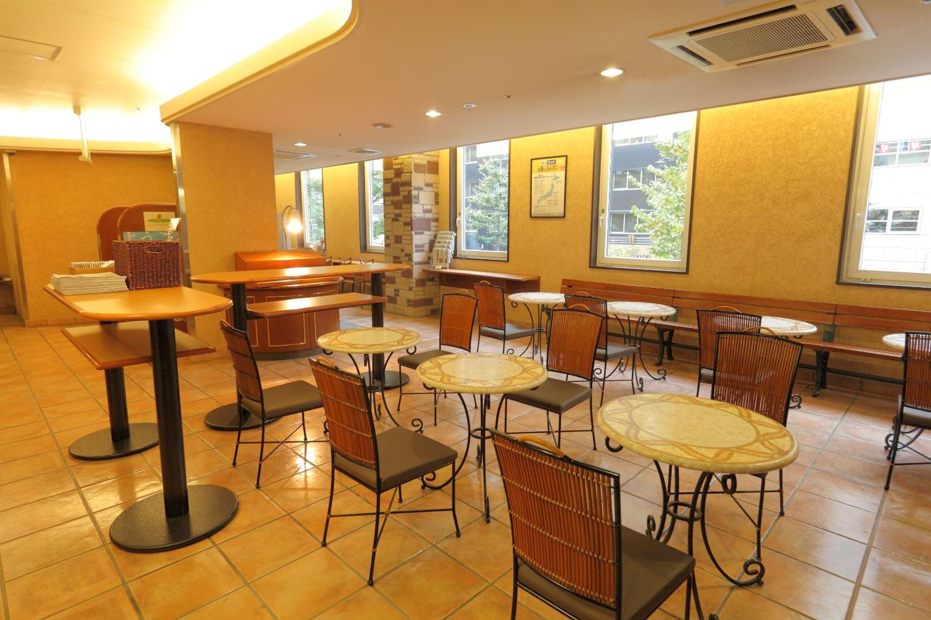 A spacious indoor area with multiple seating arrangements including tables and chairs, large windows providing natural light, and a tiled floor.