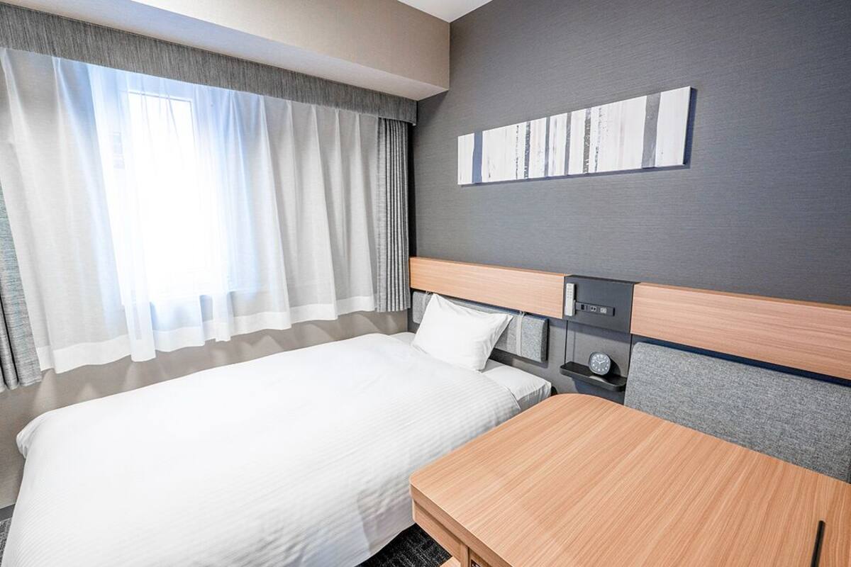 Una habitación de hotel con una cama, un escritorio de madera, una mesita de noche, un reloj y una obra de arte colgada en la pared.