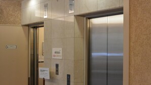 Un ascenseur avec un intérieur métallique et une porte en verre. Des panneaux sur le mur indiquent le sens de la marche de l’ascenseur et un symbole représentant l’accessibilité pour les personnes en fauteuil roulant.