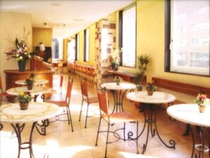 Un pasillo de hotel con mesas y sillas redondas, plantas en macetas y una larga zona con bancos.