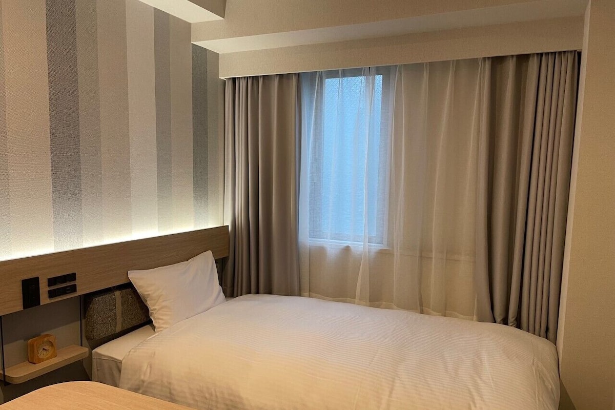 A hotel room with a bed, a bedside table, a nightstand, and a window with curtains.