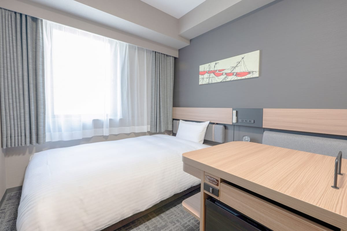 A hotel room with a bed, a wooden desk, a wall-mounted artwork, and a window with curtains.