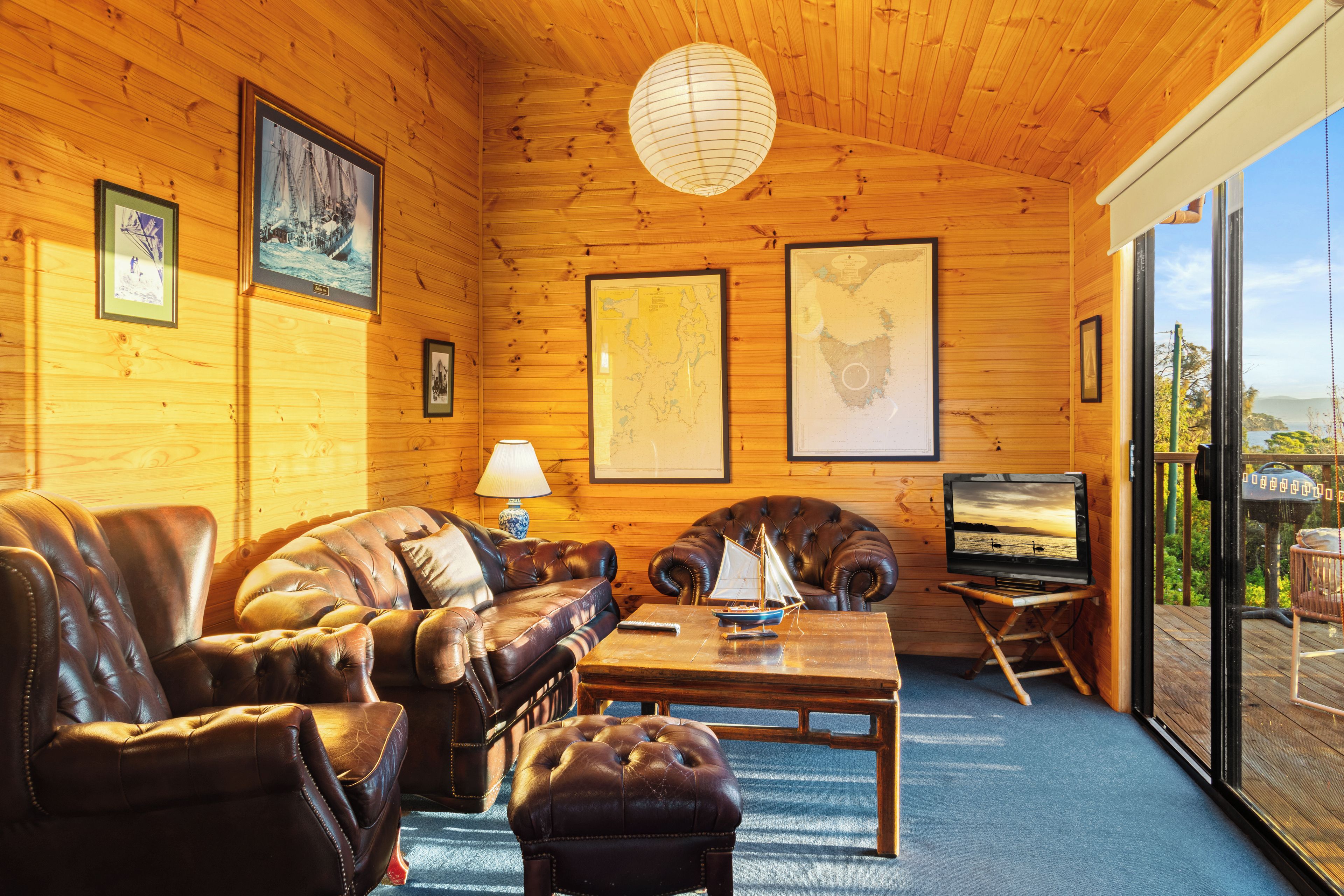 A wooden cabin interior with a leather sofa, a television on a stand, and framed maps on the walls.