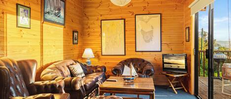 A wooden cabin interior with a leather sofa, a television on a stand, and framed maps on the walls.